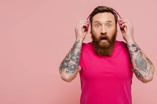 Uomo tatuato sorpreso con la bocca aperta che tiene cuffie wireless mentre guarda la fotocamera isolata sul rosa — Foto stock