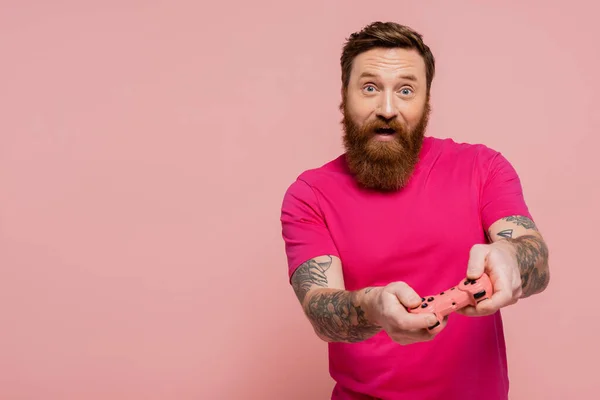 КИЕВ, УКРАИНА - 12 ДЕКАБРЯ 2022 года: взволнованный бородатый мужчина в пурпурной футболке играет в видеоигру, изолированную на розовом — стоковое фото
