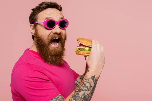 Hombre emocionado en gafas de sol de moda que abren la boca cerca de sabrosa hamburguesa aislada en rosa - foto de stock