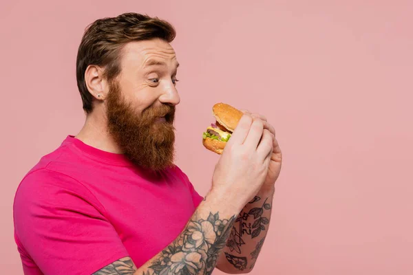 Довольный бородатый мужчина в пурпурной футболке с вкусным гамбургером, изолированным на розовом — стоковое фото