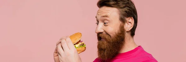 Hombre barbudo emocionado mirando deliciosa hamburguesa aislada en rosa, bandera - foto de stock