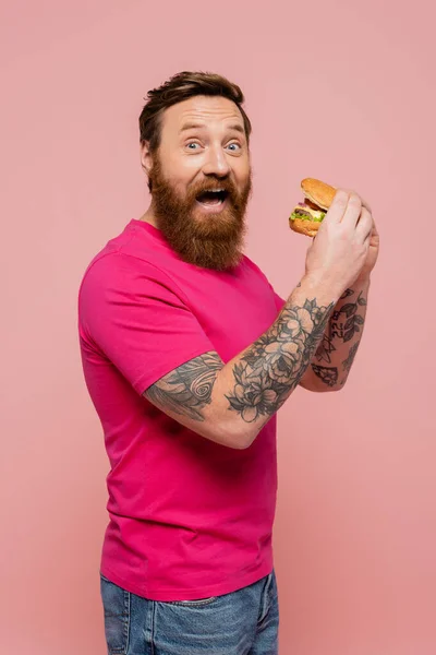 Hombre tatuado excitado en camiseta brillante mirando a la cámara mientras sostiene hamburguesa aislada en rosa - foto de stock