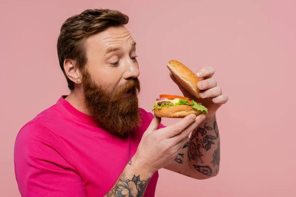 Erstaunt bärtiger Mann schaut leckeren Hamburger isoliert auf rosa — Stockfoto
