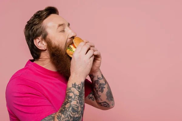 Hombre barbudo y tatuado en camiseta magenta comiendo deliciosa hamburguesa aislada en rosa - foto de stock
