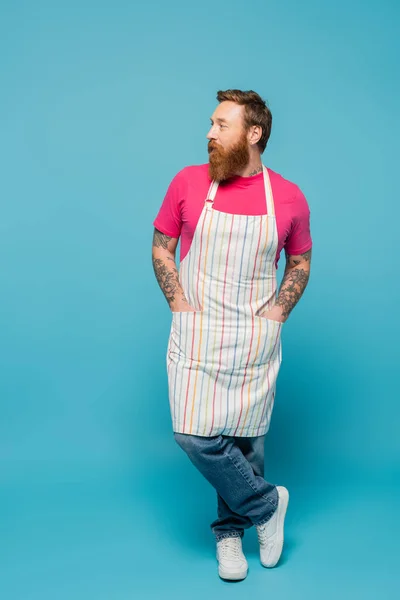 Full length of tattooed man standing with hands in pockets of striped apron and looking away on blue background - foto de stock