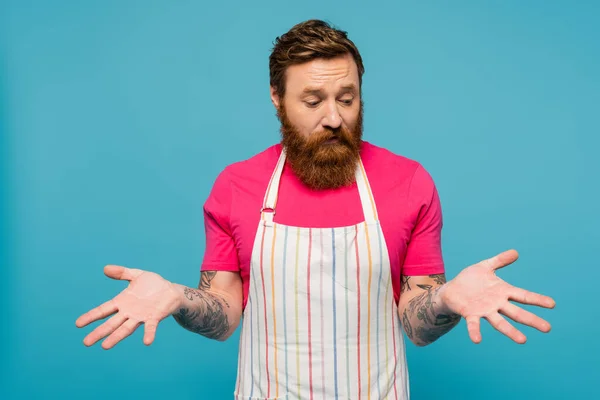 Upset bearded man in striped apron looking at empty hands isolated on blue — Stock Photo