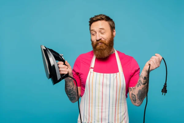 Skeptical bearded man in striped apron looking at iron and smiling isolated on blue - foto de stock