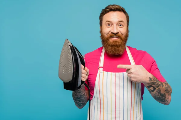 Happy tattooed man in striped apron looking at camera while pointing at iron isolated on blue — Stock Photo