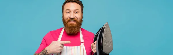 Cheerful bearded man in apron pointing at iron and looking at camera isolated on blue, banner — Stockfoto