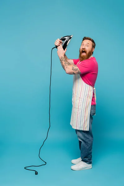 Full length of thrilled man with iron shouting and looking at camera on blue background — Stock Photo