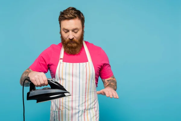 Bearded man in magenta t-shirt and striped apron holding iron isolated on blue — Stock Photo