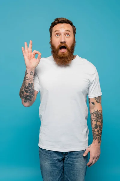 Impressed bearded man in white t-shirt looking at camera and showing okay sign isolated on blue — Stockfoto