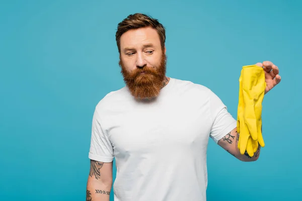 Displeased bearded man in white t-shirt holding yellow rubber gloves isolated on blue — Stockfoto