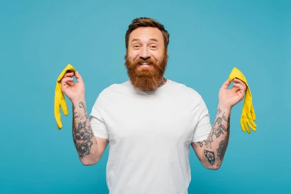 Joyful tattooed man in white t-shirt looking at camera while holding yellow rubber gloves isolated on blue - foto de stock