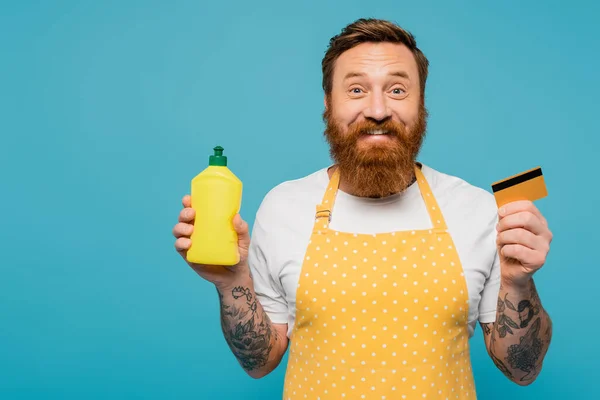 Cheerful tattooed man in yellow apron showing detergent and credit card isolated on blue — Stock Photo