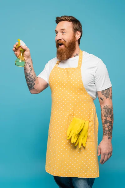 Здивований татуйований чоловік в жовтому фартусі, що тримає пляшку розпилювача ізольовано на синьому — стокове фото
