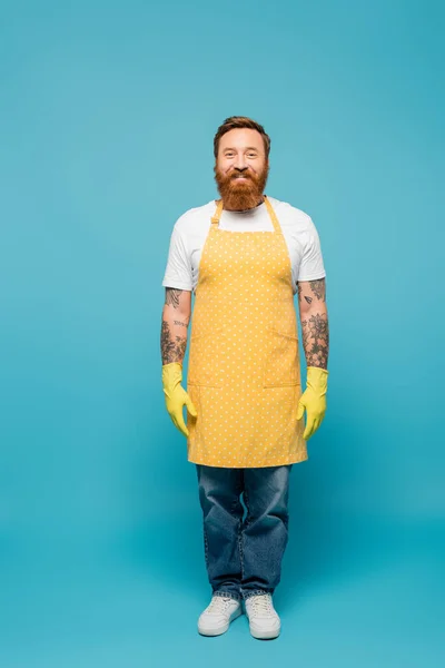 Vollbärtiger tätowierter Mann in gelber Schürze und Gummihandschuhen, der vor blauem Hintergrund in die Kamera lächelt — Stockfoto