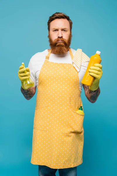 Displeased man in yellow apron puffing cheeks while standing with detergent and spray bottle isolated on blue — Fotografia de Stock