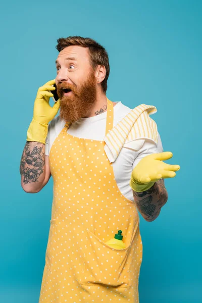 Excité homme barbu à pois tablier parlant sur smartphone et pointant avec la main isolé sur bleu — Photo de stock