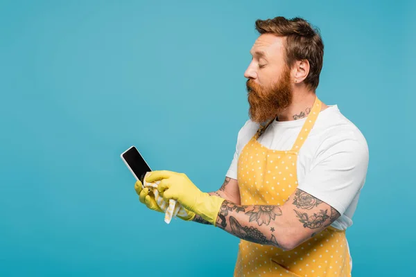 Bearded man in white t-shirt and yellow apron wiping smartphone with blank screen isolated on blue — Fotografia de Stock