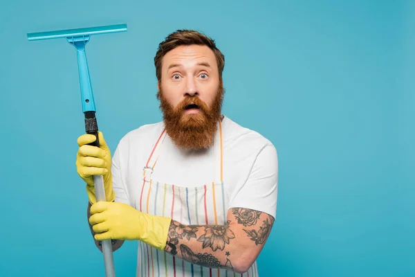 Aufgeregter Mann in weißem T-Shirt und gestreifter Schürze hält Fensterschaber in der Hand und blickt vereinzelt in die Kamera auf blauem Grund — Stockfoto