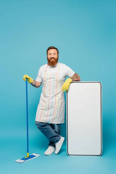 Hombre tatuado alegre en guantes de goma amarilla y delantal rayado sosteniendo trapeador cerca de plantilla de teléfono inteligente enorme sobre fondo azul - foto de stock