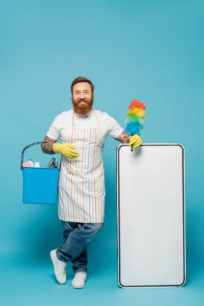 Cheerful bearded man in striped apron standing with dust brush and bucket near white phone template on blue background - foto de stock