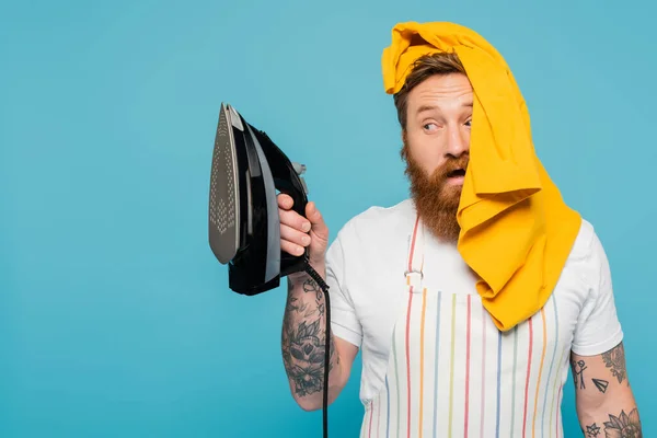 Bearded man with yellow garment on head looking at electric iron isolated on blue — Stockfoto