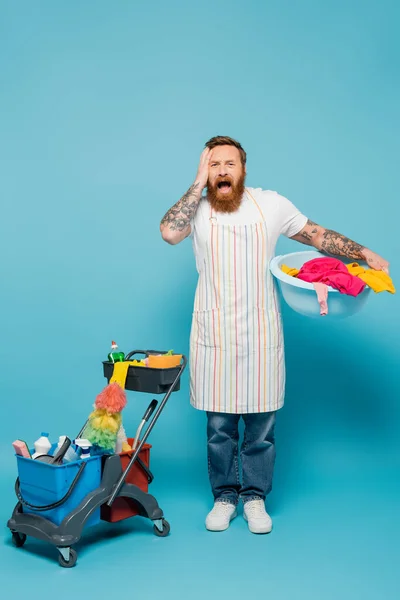 Shocked man with laundry bowl touching head and screaming near cart with cleaning supplies on blue background — Stockfoto
