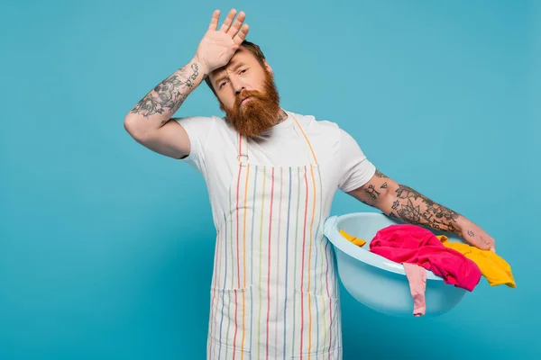 Exhausted bearded man holding hand near forehead while standing with laundry bowl isolated on blue — Stockfoto