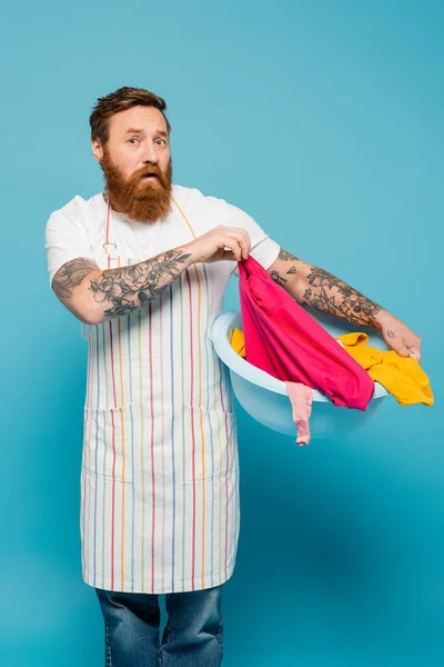 Discouraged bearded man in apron holding laundry bowl and looking at camera isolated on blue — Stock Photo