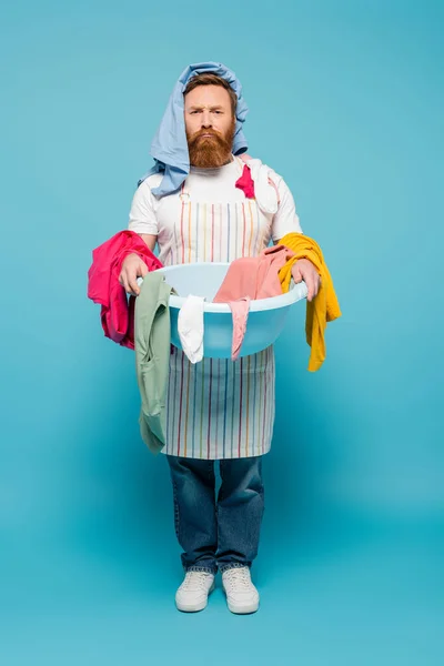 Homme barbu mécontent avec des vêtements sur la tête tenant bol à linge et regardant la caméra sur fond bleu — Photo de stock