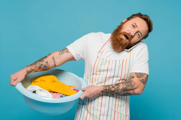 Exhausted tattooed man talking on mobile phone while holding laundry bowl isolated on blue — Stock Photo