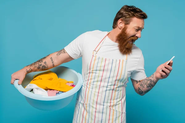 Shocked man in apron screaming while holding laundry bowl and looking at smartphone isolated on blue — Stock Photo