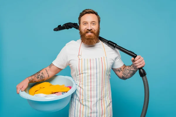 Cheerful bearded man with laundry bowl and vacuum cleaner looking at camera isolated on blue — Stock Photo