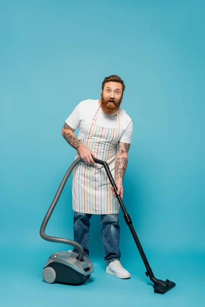 Amazed tattooed man in striped apron cleaning floor with vacuum cleaner and looking at camera on blue background — Stock Photo