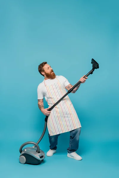 Full length of excited bearded man in striped apron having fun with vacuum cleaner on blue background — Stockfoto