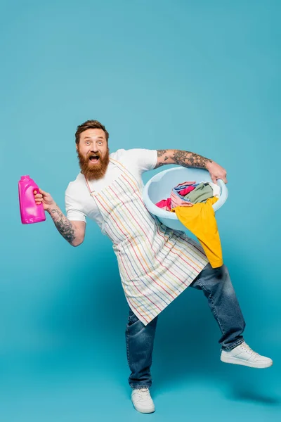 Pleine longueur de l'homme barbu excité posant avec gel de lavage et bol à linge sur fond bleu — Photo de stock