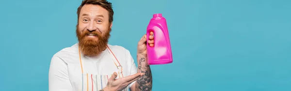 Happy bearded man in apron pointing at bottle with washing gel isolated on blue, banner — Stock Photo