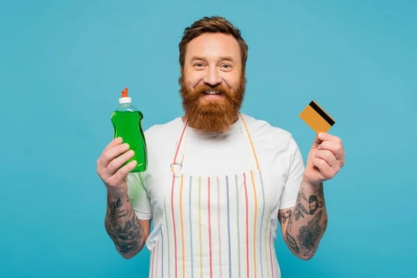 Happy bearded man with dishwashing gel and credit card smiling at camera isolated on blue — Stockfoto