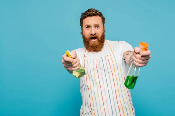 Excited man in apron having fun while spraying detergents at camera isolated on blue — Stock Photo