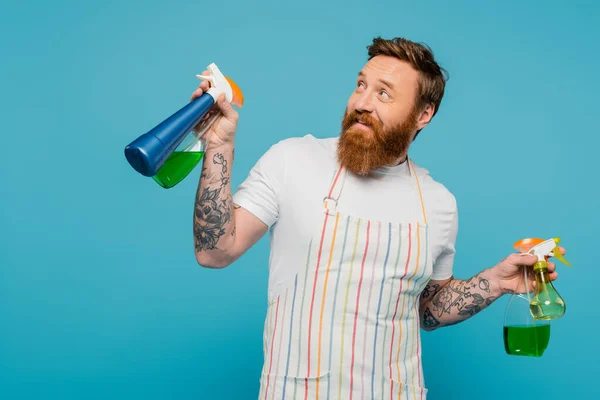 Fröhlicher bärtiger Mann mit Sprühflaschen, der lächelt und isoliert auf blauem Grund wegschaut — Stockfoto