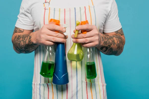 Cropped view of tattooed man in striped apron holding different spray bottles isolated on blue — Stockfoto
