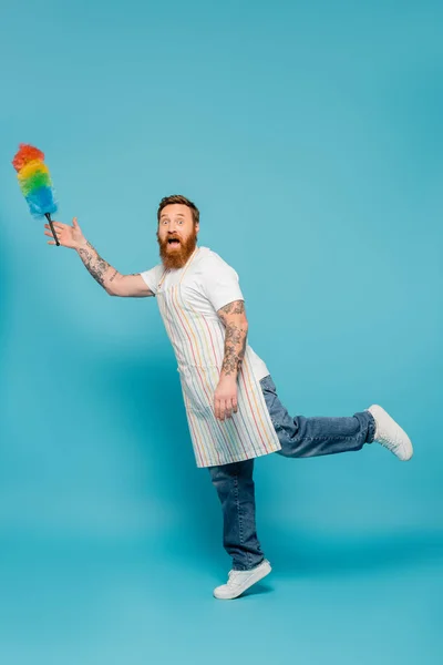 Full length of amazed bearded man in striped apron looking at camera near levitating dust brush on blue background — Stockfoto