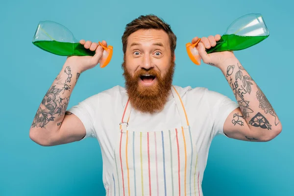 Astonished man in striped apron holding spray bottles while looking at camera isolated on blue — Stockfoto