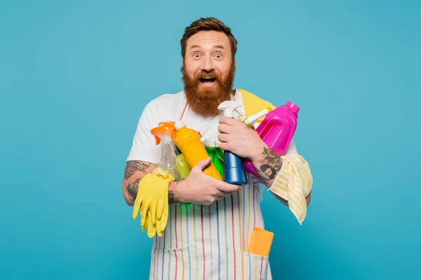 Amazed and happy man holding various detergents and rubber gloves while looking at camera isolated on blue — Stockfoto