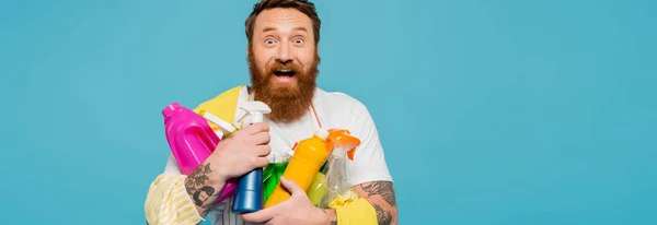 Erstaunt bärtiger Mann mit verschiedenen Waschmitteln, der isoliert auf blauem Banner in die Kamera blickt — Stockfoto