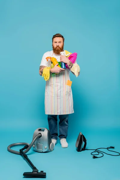 Full length of strict man holding various cleansers near iron and vacuum cleaner on blue background — Fotografia de Stock