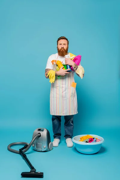 Full length of serious man holding detergents near vacuum cleaner and iron on blue background — Stockfoto