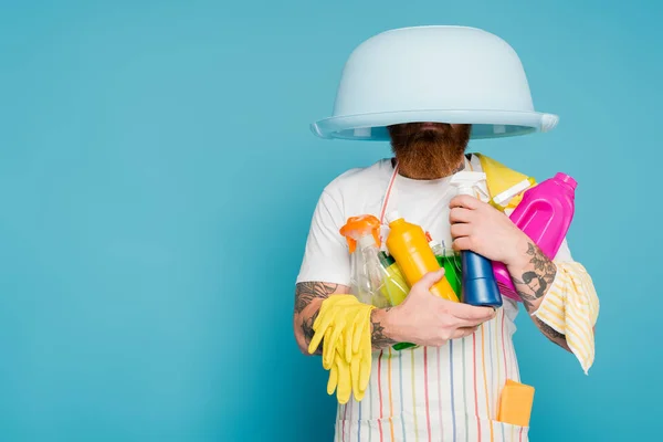 Bärtiger Mann mit verschiedenen Reinigungsmitteln und Wäscheschale auf dem Kopf isoliert auf blauem Grund — Stockfoto